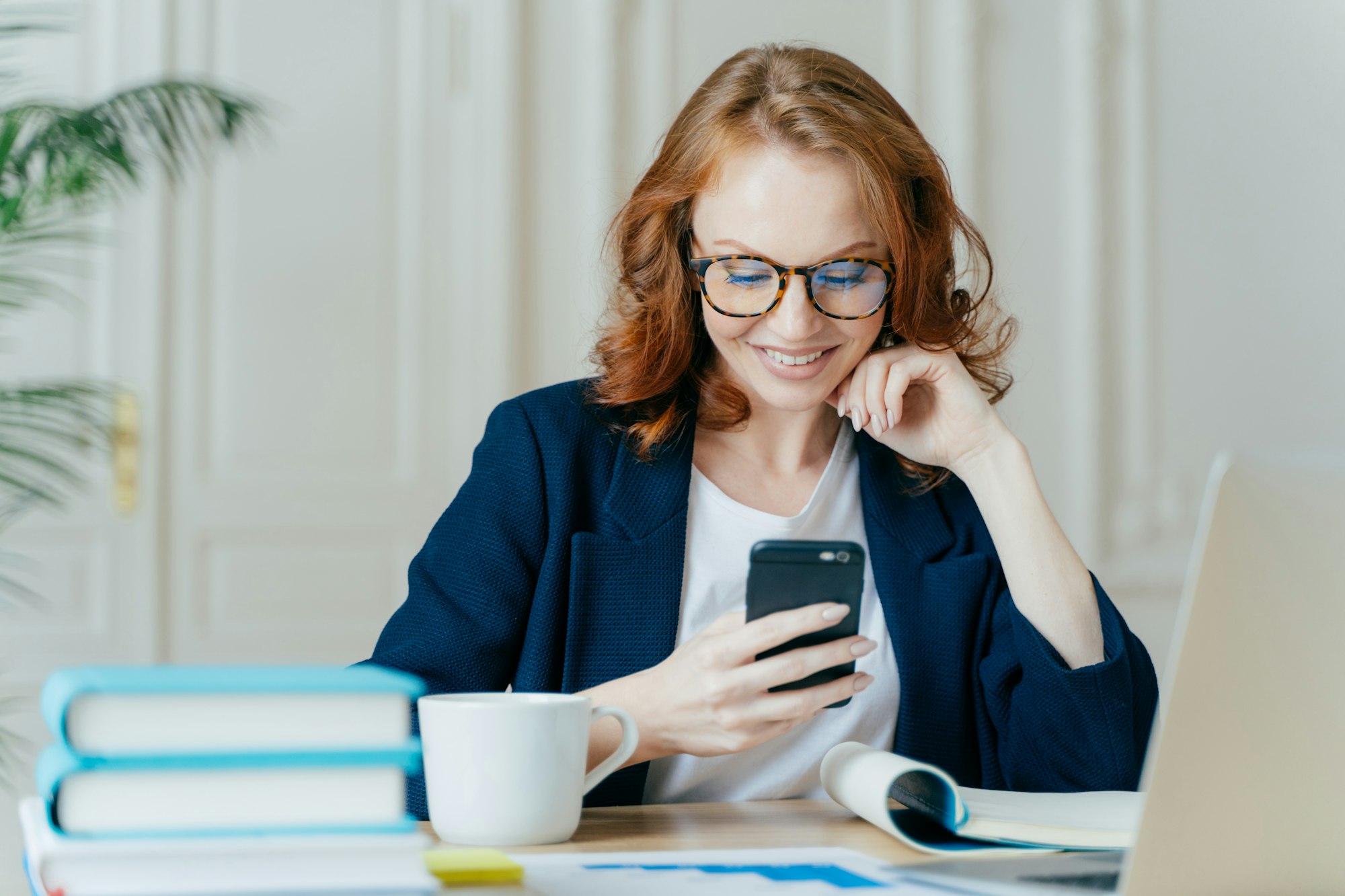 female sits with smartphone device, types feedback, works in office on up to date laptop